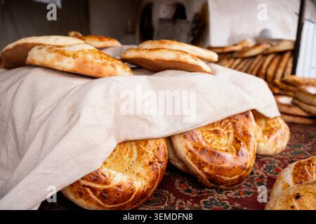 Frisch gebackene usbekische Nono- oder Lepeshka-Fladenbrote ruhen unter einem weißen Tuch in einer Bäckerei, die goldene Kruste und traditionelles Muster zeigt, Zentralasien Stockfoto