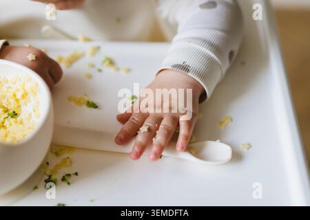 Eine Nahaufnahme der Hände eines Babys, die eine Schüssel mit kostenlosen Lebensmitteln auf einem Hochstuhl erkunden. Die Szene fängt die Neugier und Unordnung des Säuglings ein Stockfoto
