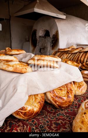 Frisch gebackene usbekische Nono- oder Lepeshka-Fladenbrote ruhen unter einem weißen Tuch in einer Bäckerei, die goldene Kruste und traditionelles Muster zeigt, Zentralasien Stockfoto