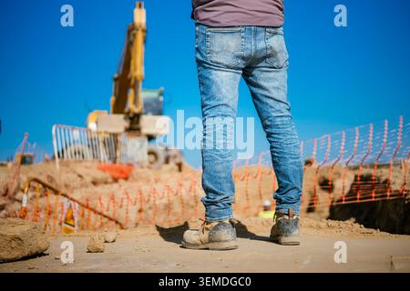 Rückansicht eines Bauarbeiters in Jeans und Sicherheitsstiefeln, der auf einer Baustelle steht, mit einem Bagger und Sicherheitszaun hinter einem durchsichtigen bl Stockfoto
