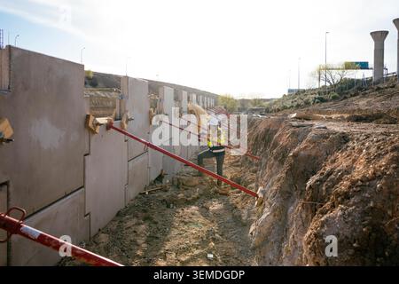 Bauarbeiterin in Schutzhelm und reflektierender Weste inspiziert Betonstützen-Wandplatten mit Streben bei einer Ausgrabung am Straßenrand unter bri Stockfoto