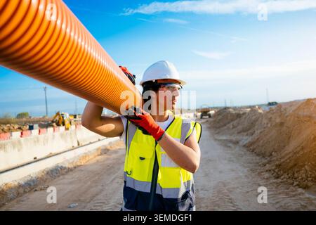 Eine Bauarbeiterin in Schutzhelm und Sicherheitsweste trägt auf einer aktiven Baustelle unter klarem blauen Himmel ein großes oranges Rohr auf der Schulter. Stockfoto