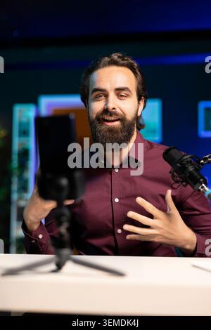 Online-Star spricht vor dem Smartphone, das im Studio am Stativstabilisator befestigt ist, und diskutiert mit den Zuschauern. Mann, der ein Mobiltelefon benutzt, um Videos im Wohnzimmer für soziale Medien aufzunehmen Stockfoto