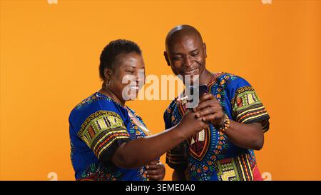 Glückliche afroamerikanische Menschen, die Webseiten auf dem Smartphone scrollen, Bilder in der Social-Media-App überprüfen und Spaß mit Memes haben. Verheiratetes Paar in traditioneller Kleidung, das Fotos online ansieht. Stockfoto