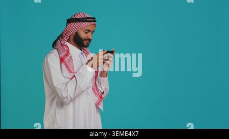Islamischer Typ, der lustige mobile Videospiele am Telefon spielt, in traditionellem Kleid mit Kopftuch gekleidet. Person aus dem Nahen Osten, die mit der Smartphone-App an Online-Gaming-Wettbewerben teilnimmt. Stockfoto