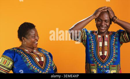 Fröhliche afroamerikanische Leute, die über etwas im Studio lachen, Spaß mit Witzen haben und sich über orangefarbenen Hintergrund amüsieren. Verheiratetes Paar lacht vor der Kamera, dumme Partner. Stockfoto