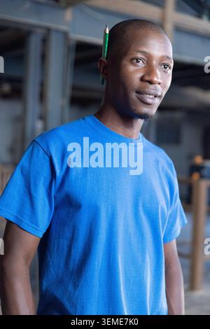 Mittelgroßer afroamerikanischer Mann, der in der Werkstatt steht, in blauem Hemd, mit grünem Bleistift hinter dem Ohr Stockfoto