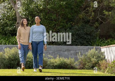 Verschiedene Freundinnen gehen Hand in Hand in Hand in einem privaten Garten in der Nähe der Pflanzwand mit Uhr Stockfoto