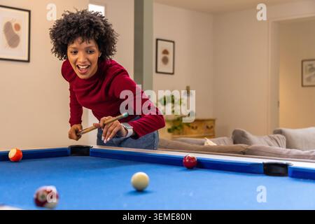 Afroamerikanerin mit rotem Oberteil, lehnt sich zu Hause über einen Billardtisch aus blauem Filz und hält Queue Stockfoto