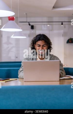 Indischer Mann in seinen Zwanzigern, der eine leichte Jacke trägt, sitzt am Tisch im Co-Working-Raum mit Laptop Stockfoto
