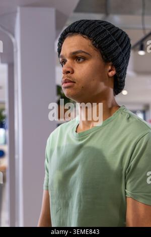 Afroamerikanischer Teenager, der in der Nähe der weißen Säule im Café steht, hellgrünes T-Shirt und dunkle Beanie trägt Stockfoto