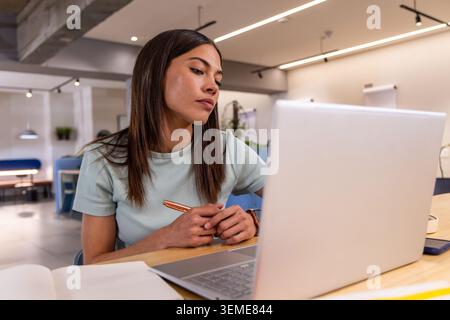 Frau, die am Tisch in der Coworking Lounge sitzt, an silbernem Laptop mit Kupferstift und Notizbuch arbeitet Stockfoto