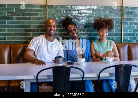 Drei Freunde sitzen auf brauner Lederbank im Café, halten Kaffeetassen und Smartphone Stockfoto