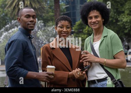 Afroamerikanische Freunde stehen am Brunnen auf der plaza in lässiger Kleidung, halten Kaffeetassen, Telefon Stockfoto