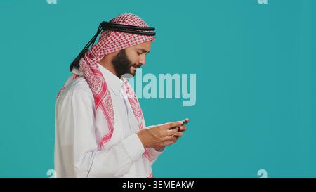 Islamischer Kerl in traditioneller Kleidung, der Online-Spiele auf dem Smartphone spielt, vor blauem Hintergrund. Arabischer Gamer, der Videospielturniere auf dem Handy genießt, Unterhaltung. Stockfoto