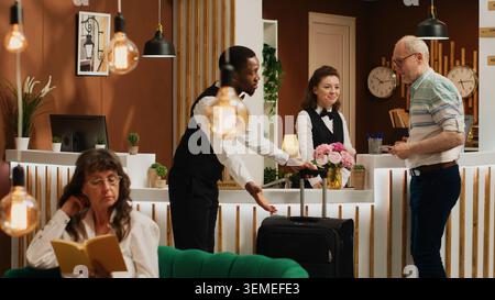 Der hilfsbereite Concierge hilft älteren Gästen beim Check-in und gibt ihnen eine Schlüsselkarte, während der Pagen sein Gepäck trägt. Rentner wartet an der Rezeption, während der Rezeptionist die Identität mit Reisepass überprüft. Stockfoto