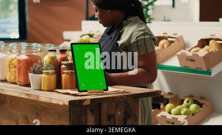 Gerät mit Greenscreen-Vorlage im lokalen Supermarkt, Tablette auf Ladentheke neben Zusatzstoffen, freie Saucen und Vorratsmaterialien in wiederverwendbaren Gläsern. Händler zeigt den Copyspace-Bildschirm an. Stockfoto