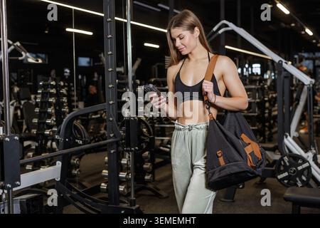 Eine fitte Frau in sportlicher Kleidung überprüft ihr Handy, während sie durch ein modernes Fitnessstudio geht und eine Mischung aus Fitness und Technologie präsentiert. Stockfoto