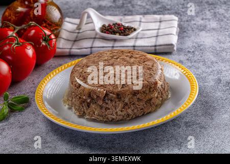 Traditionelles aspisches Fleisch-Gericht auf Keramikplatte, begleitet von frischen roten Tomaten, Basilikumblättern, gemischten Pfefferkörnern im Löffel und einer Ölflasche Stockfoto