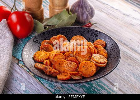 Goldene, geröstete Süßkartoffelchips in schwarzer Keramikschale, umgeben von Reifen Tomaten, Knoblauchzwiebeln, Lorbeerblättern und hölzernen Pfeffermühle auf blauem Hintergrund Stockfoto