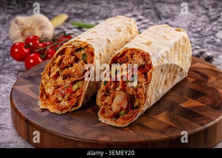Appetitliche Hähnchenhawarma-Wraps in Hälften mit saftigem Fleisch, frischem Gemüse und Gewürzen auf einem rustikalen Holzschneidebrett mit Kirschtomaten Stockfoto