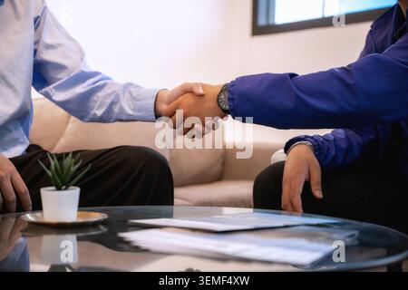 Schließen Sie Geschäftspartner, die einen Vertrag unterzeichnen, und zwei Personen schütteln sich die Hand an einem Couchtisch. Einer trägt ein hellblaues Hemd, der andere Stockfoto
