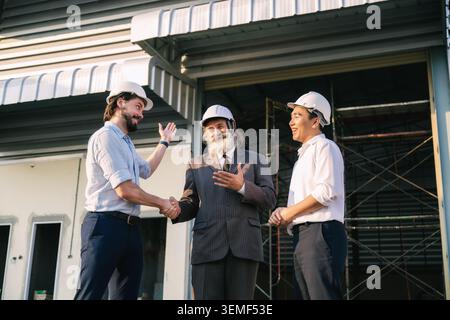 Lächelnder Geschäftsmann schüttelt die Hand Baustelleninspektion, drei Männer in weißen Schutzhüten stehen auf einer Baustelle. Man schüttelt ha Stockfoto