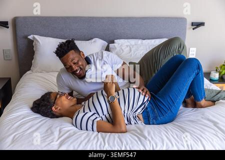 Afroamerikanisches Paar, das zu Hause auf dem Bett liegt und sich über eine schwangere Frau mit gestreiftem Oberteil beugt Stockfoto