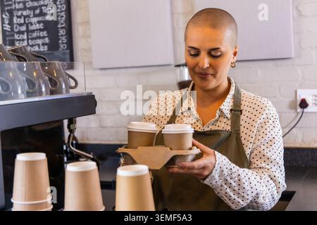 Weibliche Barista hält zwei Pappbecher in der Halterung hinter der Espressomaschine und trägt eine olivgrüne Schürze Stockfoto