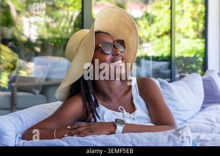 Afroamerikanerin, die sich auf einem hellgrauen Sofa in der Sonne liegend, mit Strohhut und Smartwatch Stockfoto