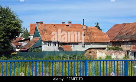 Hinter einem blauen Geländer und einer baufälligen Backsteinmauer steht ein verwittertes historisches Gebäude mit einem hängenden Dach aus Terrakotta-Fliesen und kleinen Fenstern Stockfoto
