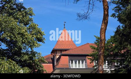Ein markanter quadratischer Pyramidendach-Turm mit orangen Terrakottafliesen und einem Metallfinial steht hinter einer Dachlinie aus Mansarden inmitten von dichtem Grün Stockfoto