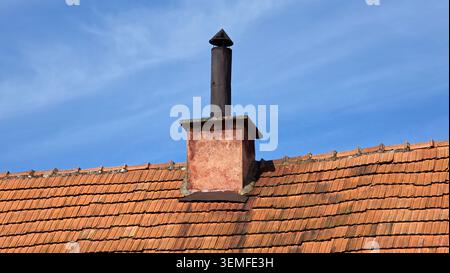 Ein quadratischer Schornsteinsockel mit Stuck trägt ein dunkles Metallrohr mit einer konischen Regenkappe auf einem Dach aus altem orangenem Ton unter blauem Himmel Stockfoto