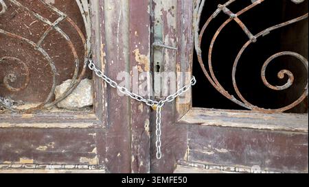 Eine Metallkette schlingt durch die Rahmen eines Paares alter brauner Türen mit abblätternder Farbe und rostigen Rollgittern, um den Eingang zu verriegeln Stockfoto