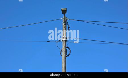 Ein verwitterter hölzerner Versorgungsmast trägt eine Straßenlaterne, Spiralkabel und horizontale Stromleitungen, die sich gegen einen blauen Himmel erstrecken Stockfoto
