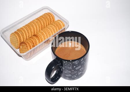 Eine Tasse heißer brauner Milchkaffee in einer schwarzen Tasse mit weißen Retro-Abbildungen, neben einem durchsichtigen Kunststoffbehälter mit runden Keksen auf einem hellweißen Hintergrund Stockfoto