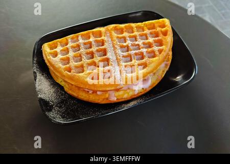 Süßes gefaltetes Waffelsandwich, gefüllt mit rosa Erdbeercreme und mit Puderzucker bestreut, serviert auf einem schwarzen Teller auf einem dunklen strukturierten Hintergrund. Lebensmittelfotografie Stockfoto