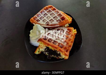 Nahaufnahme von frisch gebackenen goldenen Waffeln, gefüllt mit zerquetschten Schokoladenkeksen und Sahne, mit Puderzucker bestäubt, serviert mit weichem Vanilleeis auf einem schwarzen Teller auf dunklem Hintergrund. Stockfoto