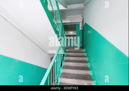 Treppenhaus mit blaugrünen und weißen Wänden. Betontreppen mit blauem Geländer führen nach oben. Oben an der Treppe ist eine Tür zu sehen Stockfoto