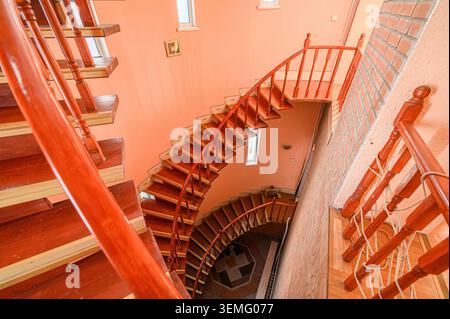 Hochwinkelaufnahme einer hölzernen Treppe mit geschwungenem Design, an pfirsichfarbenen Wänden und einem Backsteinabschnitt. Fenster bieten Tageslicht. Stockfoto