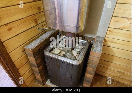 Der mit Felsen gefüllte Saunaofen befindet sich in einer Ecke einer Holzsauna. Der Herd hat einen Metallkamin und Ziegelstein. Die Wand besteht aus hellen Farben Stockfoto
