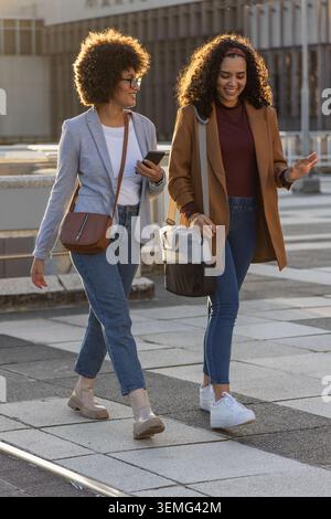 Zwei weibliche Freunde, die sich über die gekachelte plaza unterhalten, Blazer tragen und Telefon und Taschen halten Stockfoto
