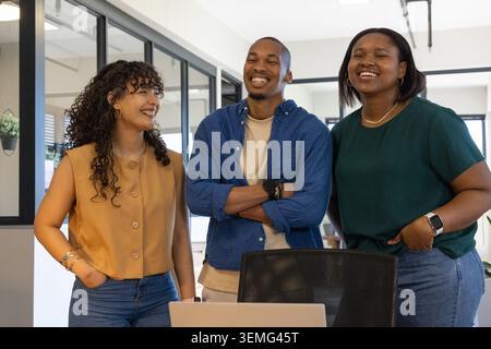 Verschiedene Kollegen lächeln und stehen am modernen Schreibtisch mit Laptop und Stuhl in der Nähe von Glaswänden Stockfoto