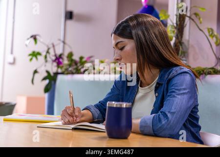 Frau, die in offenem Notizbuch mit Metallstift in der Lounge schreibt, trägt ein Denim-Hemd in der Nähe eines blauen Tumbler Stockfoto