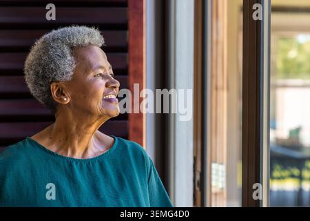 Eine ältere afroamerikanische Frau posiert vor einer Schiebetür zu Hause in der Nähe eines hölzernen Lamellen mit einer blauen Bluse Stockfoto