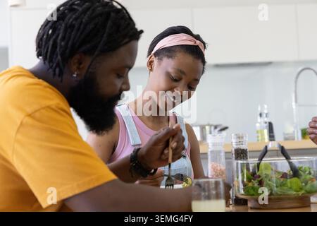 Afroamerikanisches Paar sitzt am Holztisch in der Küche, Mann in Senftee, Teen servieren Salat Stockfoto