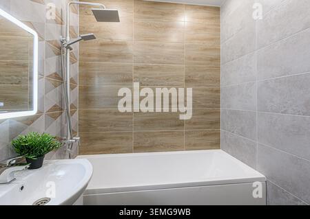 Das gut beleuchtete Badezimmer verfügt über eine weiße Badewanne, ein Waschbecken mit Pflanzen und geflieste Wände. Eine Wand hat eine holzähnliche Fliese, während die andere grau ist. Ein beleuchteter Spiegel A Stockfoto