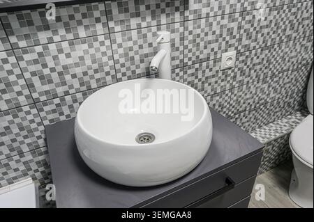 Das moderne Badezimmer verfügt über ein rundes, weißes Keramikwaschbecken mit Chromabfluss und einen eleganten weißen Wasserhahn. Das graue, geflieste Backsplash und die Kabine Stockfoto