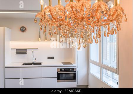 Über einer modernen, weißen Küche hängt ein eleganter bernsteinfarbener Kronleuchter. Die Küche verfügt über elegante Schränke, ein schwarzes Waschbecken und große Fenster. Stockfoto