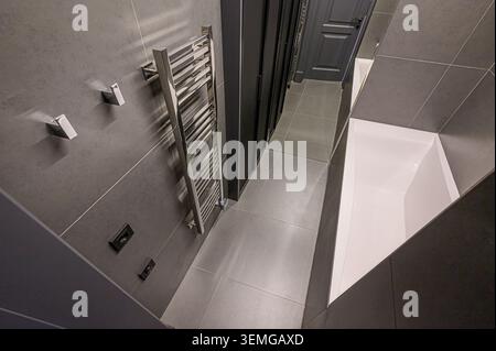 Das moderne Bad ist mit grauen gefliesten Wänden und Böden, einem Handtuchwärmer aus Chrom, Wandhaken, einem dunklen Schrank und einer weißen Badewanne ausgestattet. Das Design ist schlank und m Stockfoto